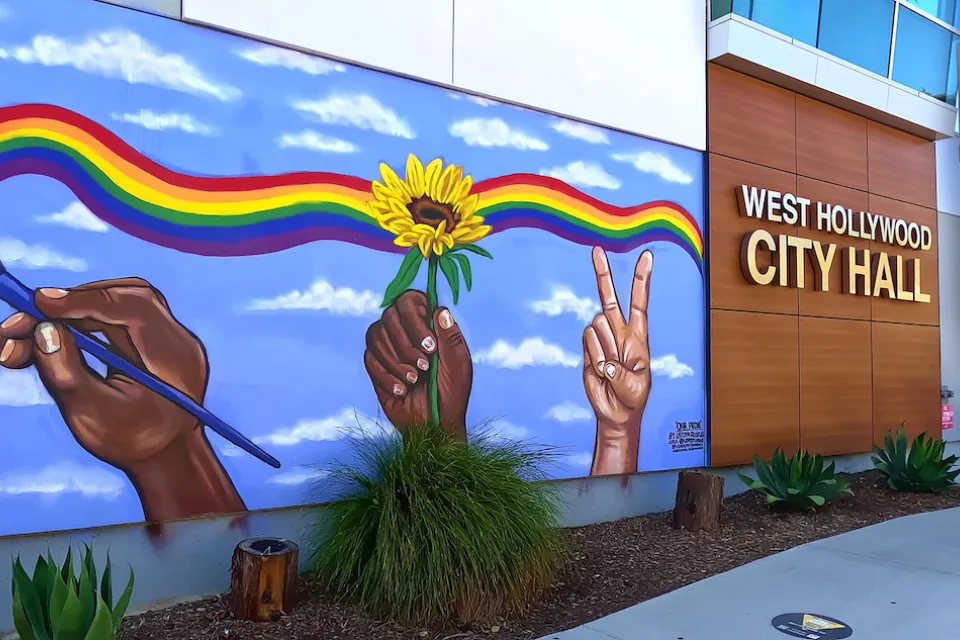 Front entrance of West Hollywood City Hall with name on side of building and mural of hands painting a rainbow across a blue sky.